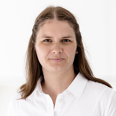Eine Frau mit langen, braunen Haaren trägt ein weißes Hemd und schaut direkt in die Kamera mit einem neutralen Ausdruck.