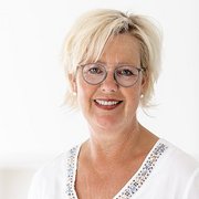 Eine lächelnde Frau mit blonden Haaren und Brille, die in einem hellen Raum steht und eine freundliche Ausstrahlung hat.