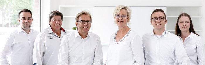 Professionelles Teamfoto in heller Büroumgebung Sechs Personen in weißen Hemden stehen in einem modernen Raum, lächeln freundlich und wirken professionell.