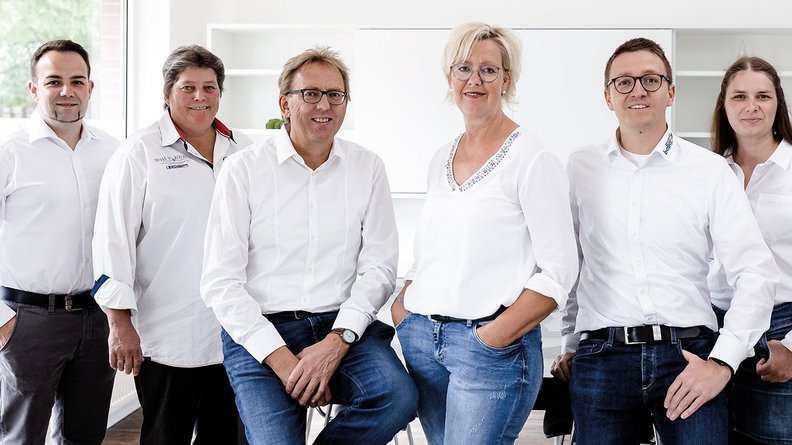 Sechs Personen in weißen Hemden stehen in einem modernen Raum, lächeln freundlich und wirken professionell.