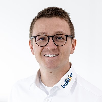 Ein junger Mann mit Brille lächelt freundlich in die Kamera, trägt ein weißes Hemd mit einem Logo auf dem Kragen.