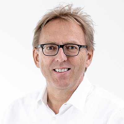 Ein lächelnder Mann mit Brille trägt ein weißes Hemd und hat eine freundliche Ausstrahlung vor einem hellen Hintergrund.