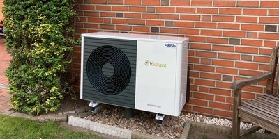 Eine moderne Wärmepumpe steht neben einer roten Ziegelwand, umgeben von einem gepflegten Garten mit Sträuchern.