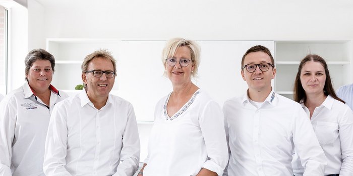 Fünf Personen in weißen Hemden stehen in einem hellen Raum, lächeln freundlich und wirken professionell.